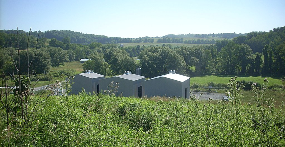A rural natural gas compressor station (three gray compressor units housed in small buildings) along a gathering system in a producing region. These field compressors boost the pressure of gas collected from nearby wells, pushing it through the network toward a processing plant.